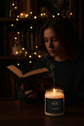 A white wax candle in a clear glass jar with a black metal lid and a black label featuring gothic-style text and illustrations.